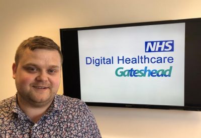Head and shoulders shot of Adam Charlton with Digital Health Gateshead on a tv in the background